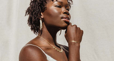 Women in sun in front of a light background with gold and wite earrings and gold necklace and bracelet