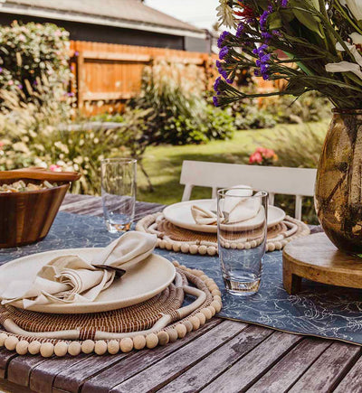 Table setting with beaded chargers (white and tan) with plates and neutral colored napkins and hexagon over rings. there is a blue table running ontop of the wooden table. This table is outside. 