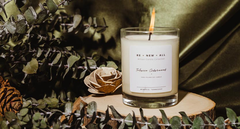 White scented candle standing on a wood pedestal in  rustic holiday greenery setting.