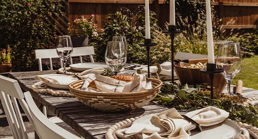 Wooden table outside with placemats, napkins, baskets, wine glasses, candle sticks