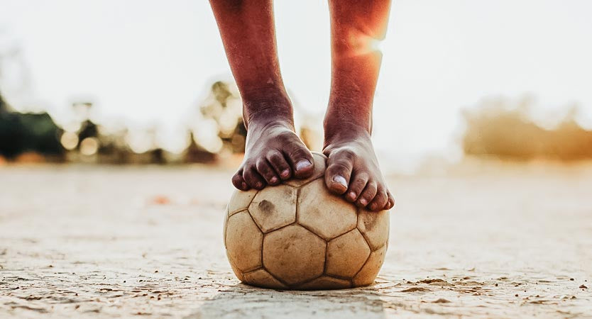Child's feet standing on top of a deflated soccer ball on the sand