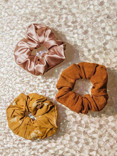 Three scrunchies on a table. One is pink, one is orange. one is yellow. 