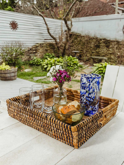 Square woven basket on white table with glasses, flowers and bread inside. 