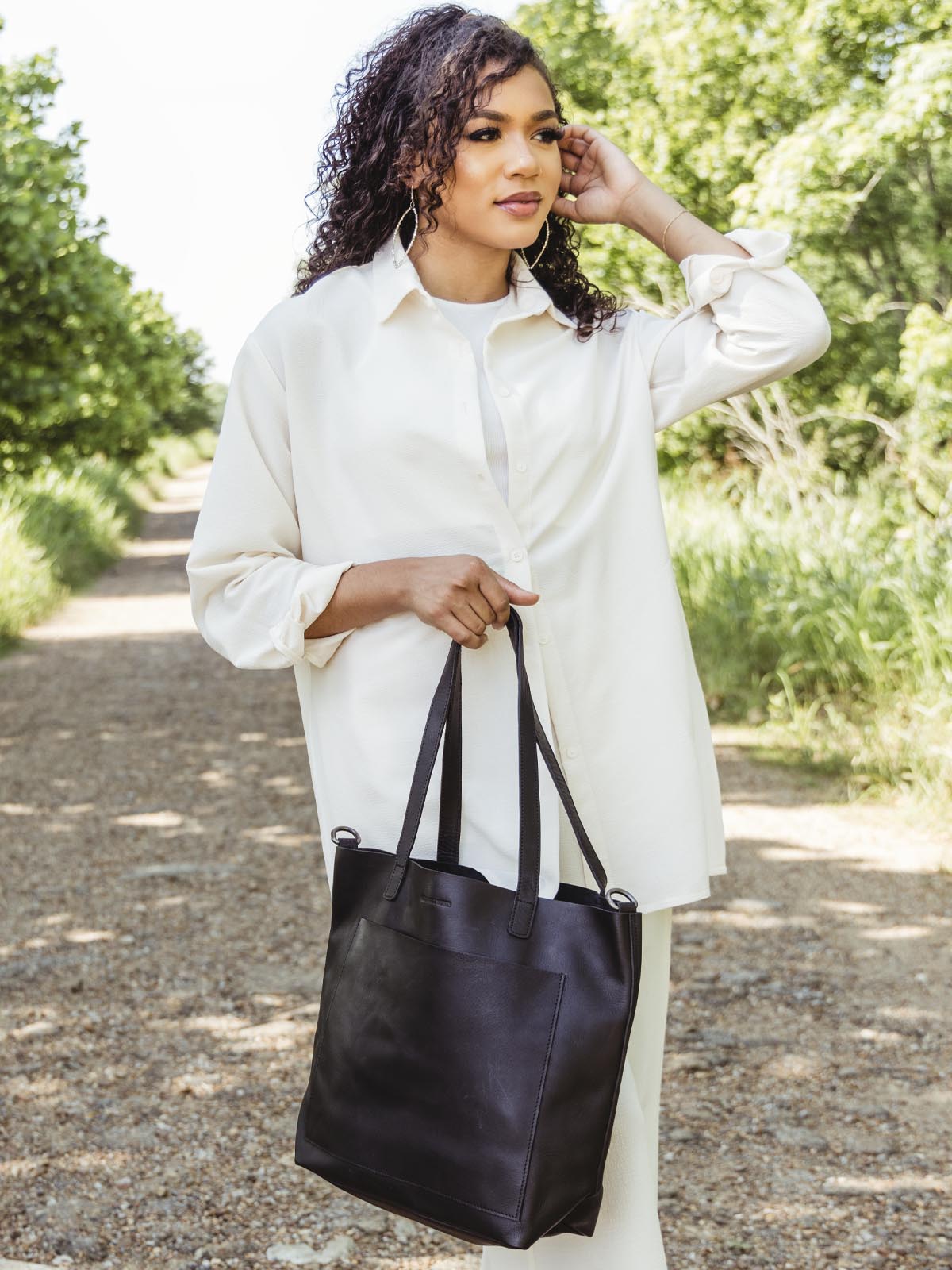 Model in an outdoor setting with made free black leather bag. The model is show holding the bag using the hand straps. 