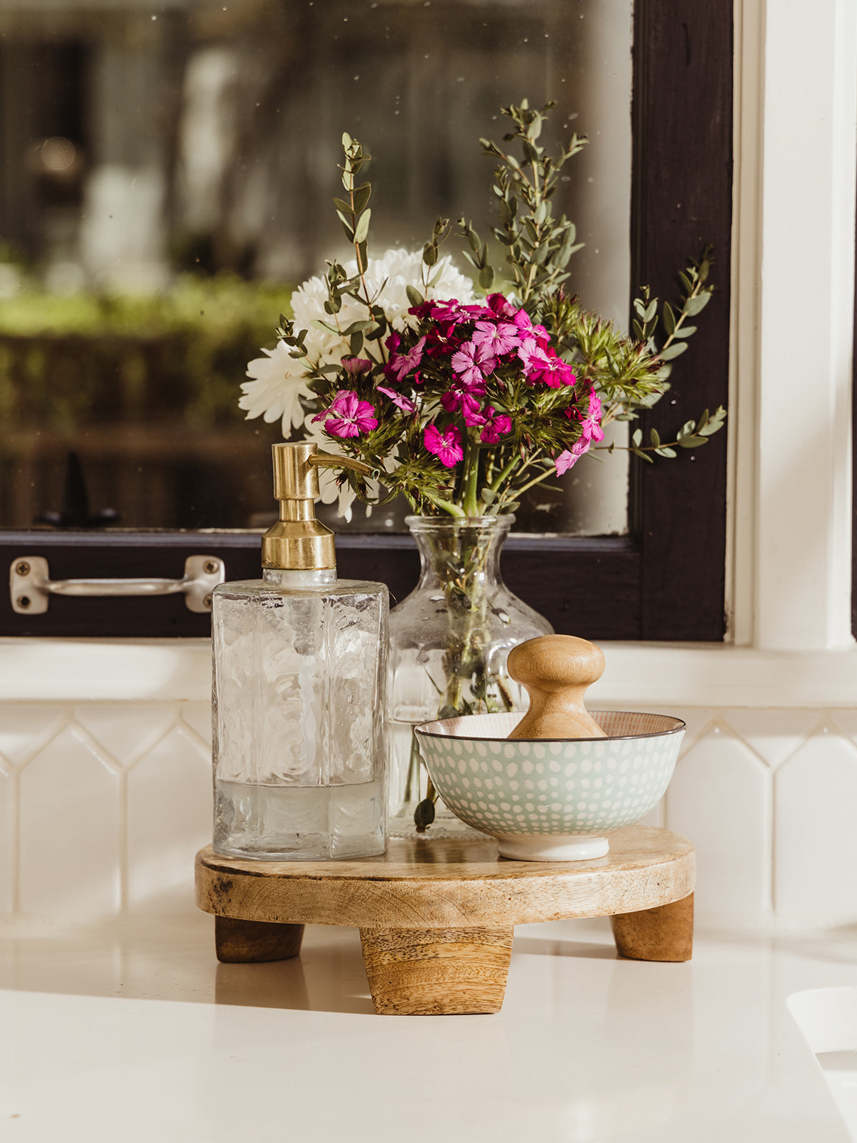 Small mango wood tray holding soap dispenser and flower vase atop. Placed on a white counter in front of a window. 