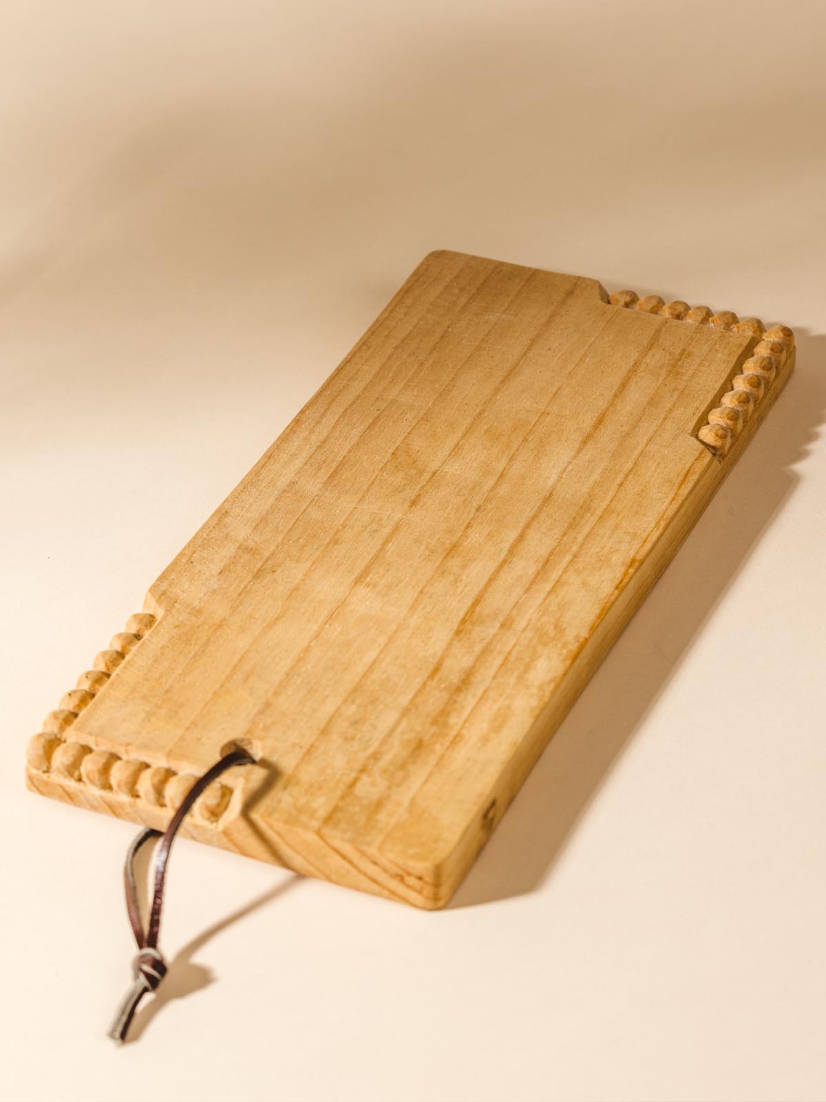 Small rectangular handmade serving board with circular detail carving on the top right and bottom left corners. Top of board contains a dark leather strap for hanging.