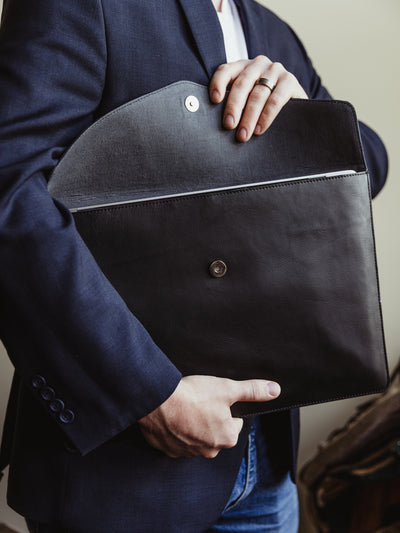 Male model opening black leather laptop sleeve with envelope style opening and metal snap. 