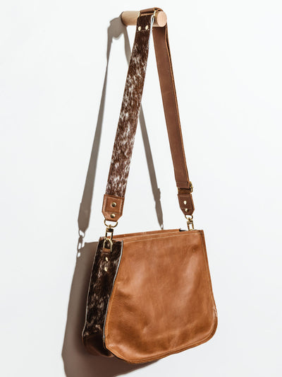 Brown Leather Messenger Bag with Cowhide hanging on white wall showing off the cowhide strap and sides of the purse. 