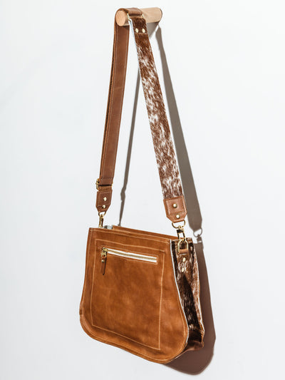 Brown Leather Messenger Bag with Cowhide  hanging on white wall showing the back side of the purse with an exterior zipper. Also visible, the cowhide strap and sides. 