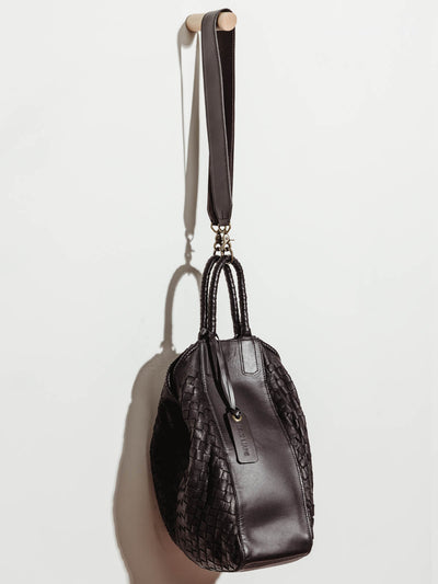 Front view of black leather all day tote closed shut, hanging by shoulder strap on white wall