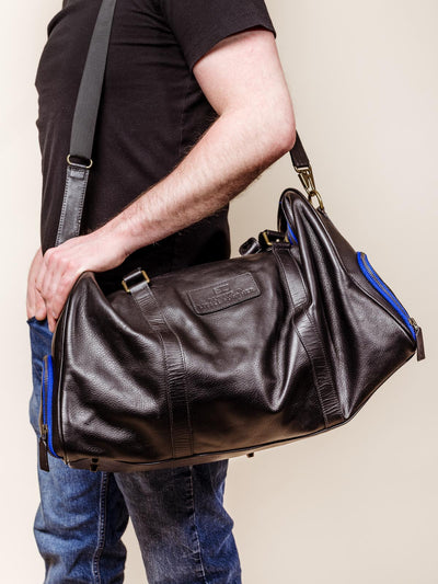 Male model holding black leather duffle bag on cream studio background.