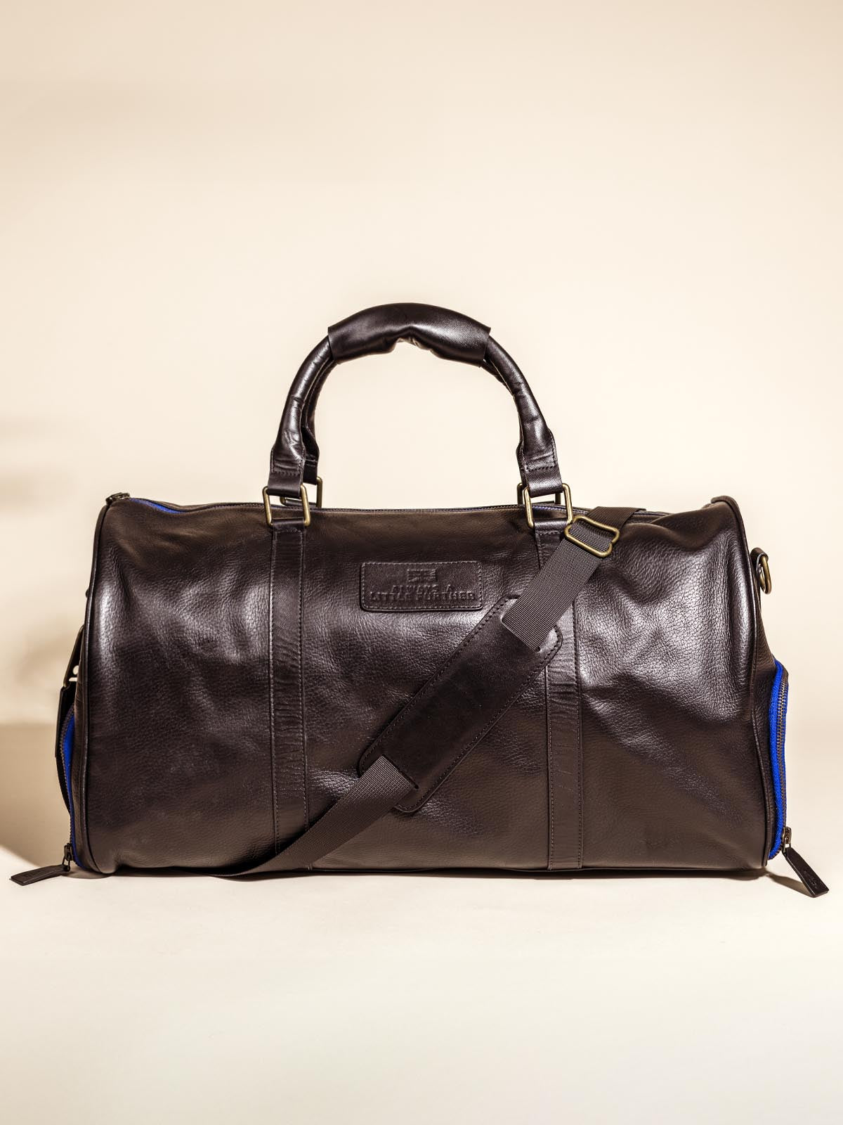 Black Leather Duffle Bag on off white backdrop featuring shoulder straps, hand straps, and blue accent zippers