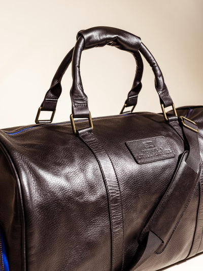 Close up angled image of the Black Leather Duffle Bag on an off white backdrop featuring shoulder straps, hand straps, and blue accent zippers