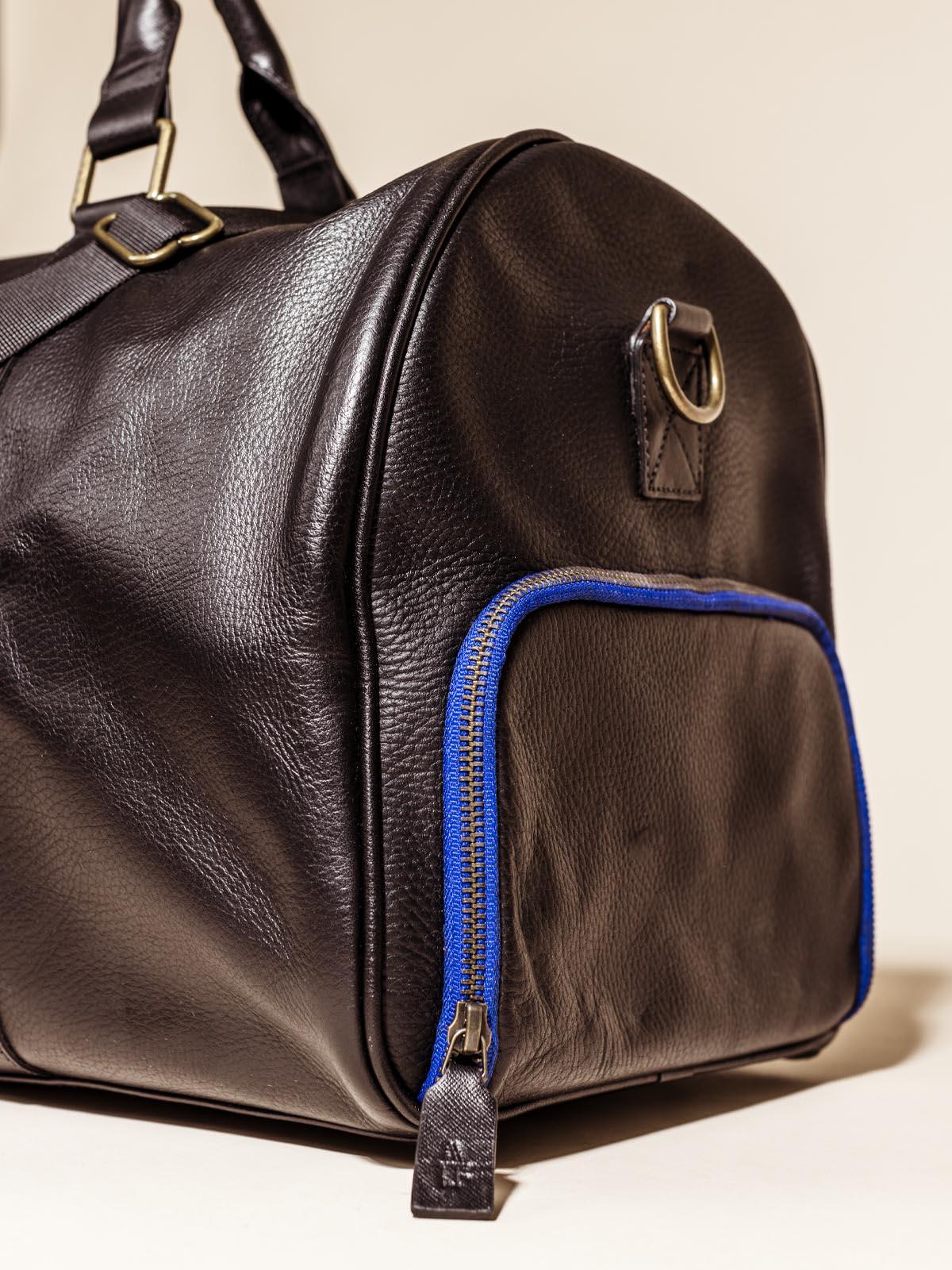 Close Up of the side of the Black Leather Duffle Bag on an off white backdrop featuring shoulder straps, hand straps, and blue accent zippers