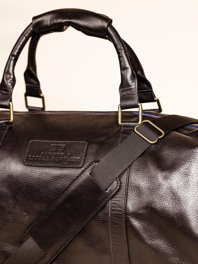Close Up of the top of the Black Leather Duffle Bag on an off white backdrop featuring shoulder straps, hand straps, and blue accent zippers
