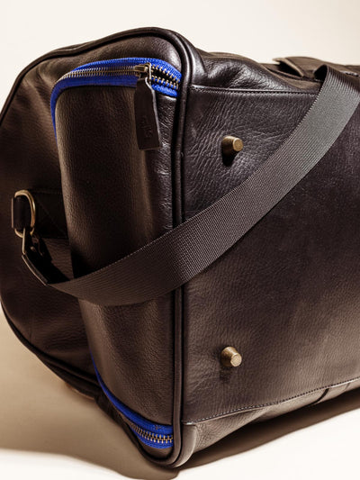 Close Up of the bottom of the Black Leather Duffle Bag on an off white backdrop featuring brass pegs, shoulder straps, and blue accent zippers
