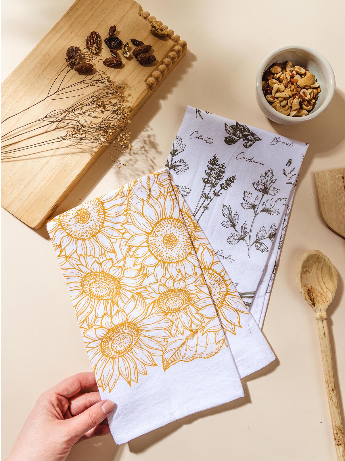 Watercolor tea towels on cream surface with hand touching the sunflower pattern tea towel. Featured: cutting board, wooden utensils, flowers, and food for styling. 
