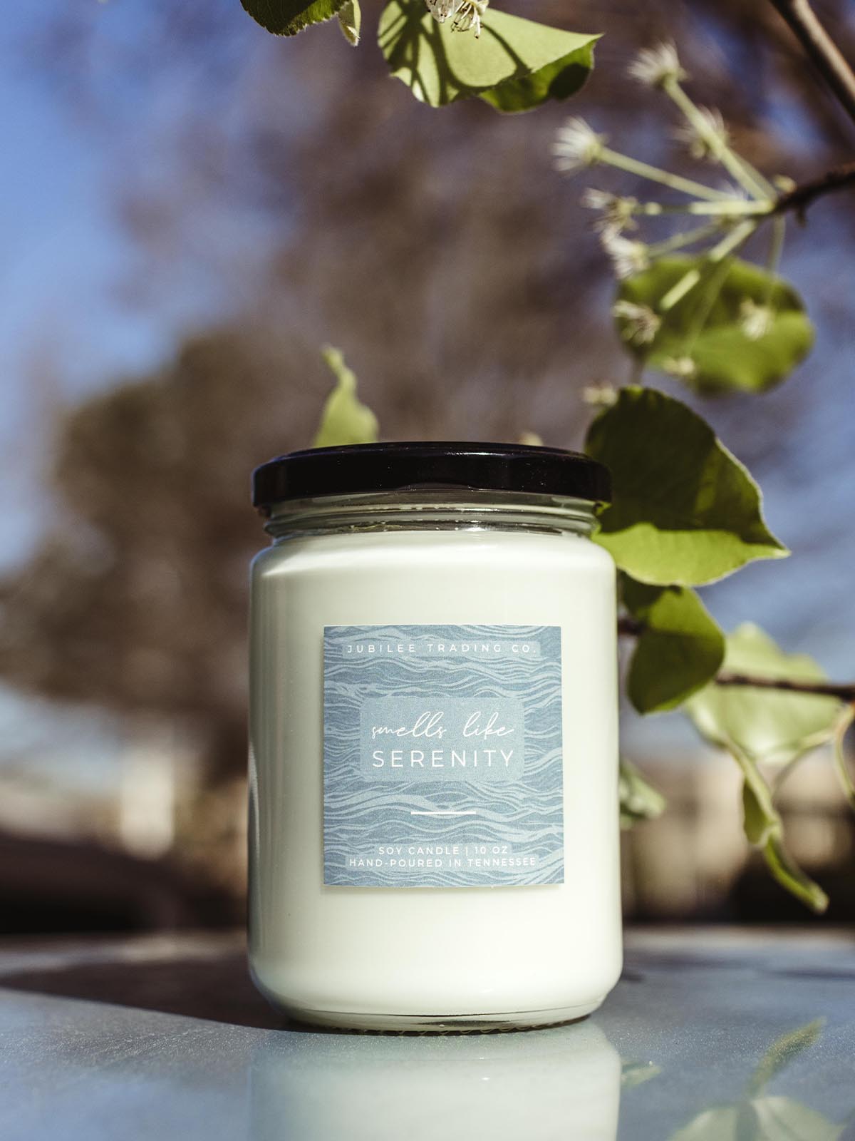 Smells Like Serenity candle on a blue surface with blue sky and greenery in the background. 