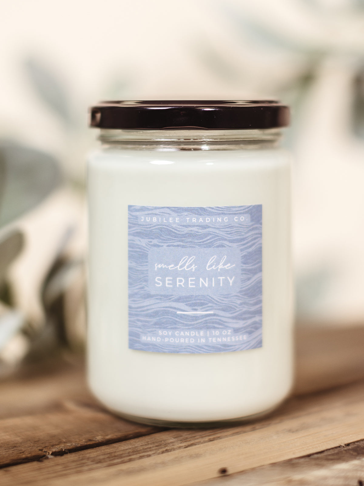 Smells Like Serenity scented candle on a wooden surface with greenery for styling. 