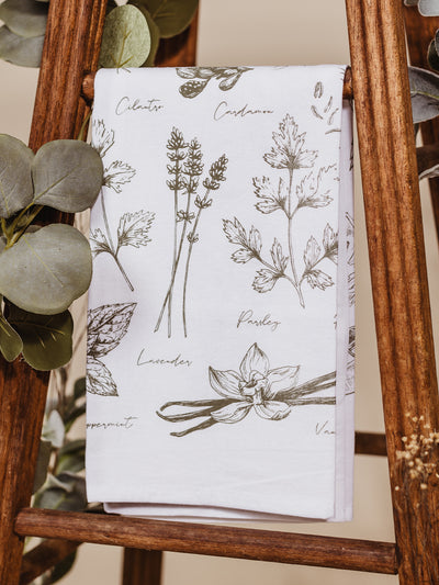 Herbs line art tea towel dropped on a wooden stool with greenery.