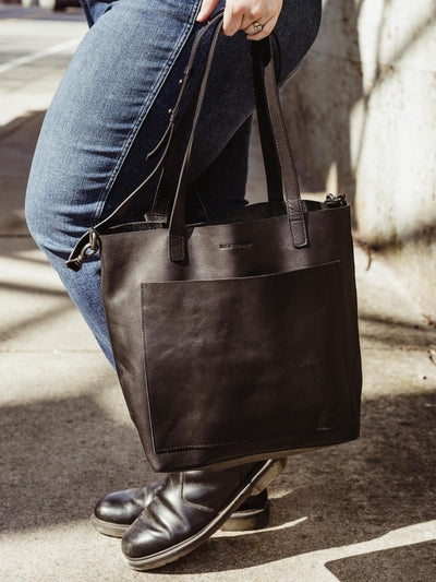 Female model holding black leather tote low to the ground outdoors.