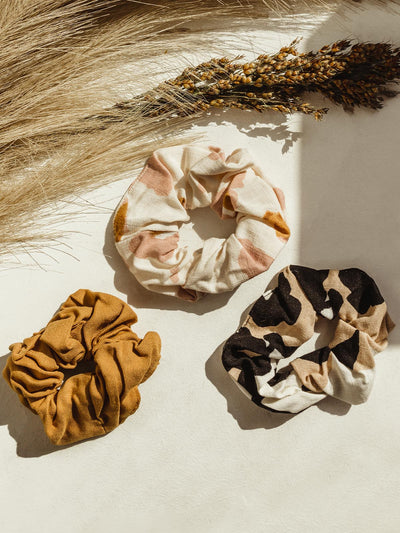 Set of three scrunchies on a cream table with flowers. One Mustard Color. One black and tan abstract print. One blush pink, mustard, and cream abstract print.