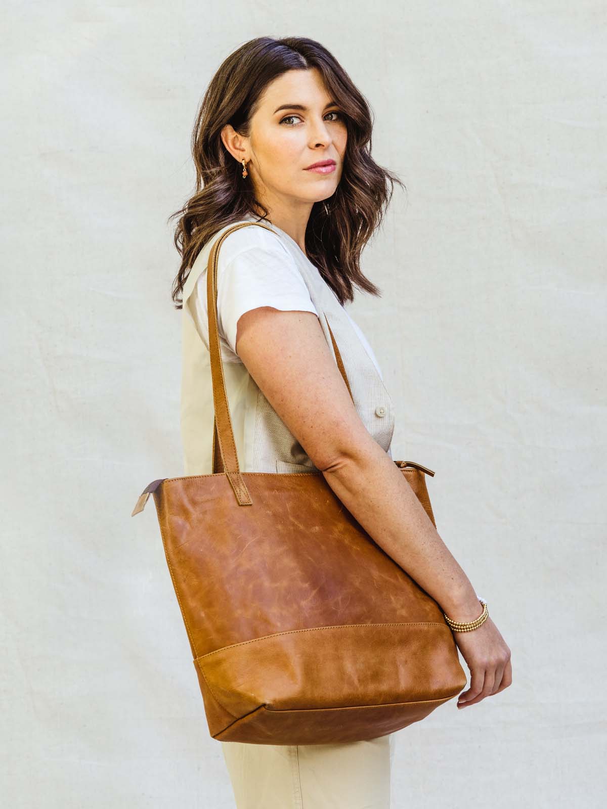 Brunette female model holding hazelnut colored leather tote bag over the shoulder. 