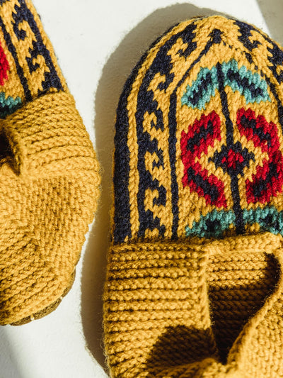 Close up of golden yellow hand-knitted mustard shoe/socks. Across the top is a tribal like pattern in red, blue-green, and navy.