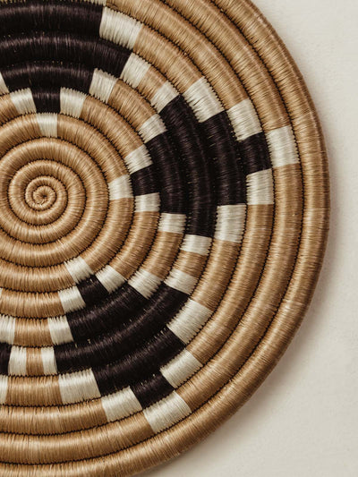 Small handwoven circular trivet on white counter. Tan base with black and white pattern.