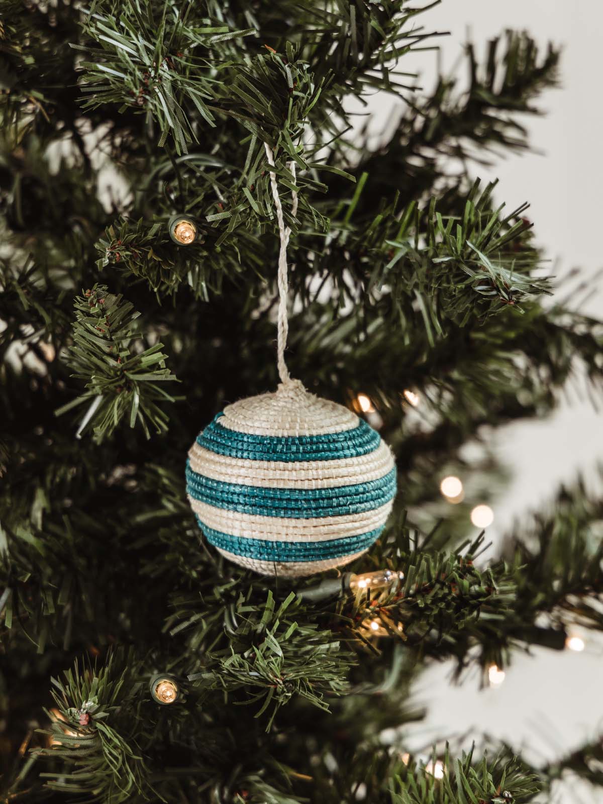 White and blue-green woven striped ball ornament hanging on holiday tree. 