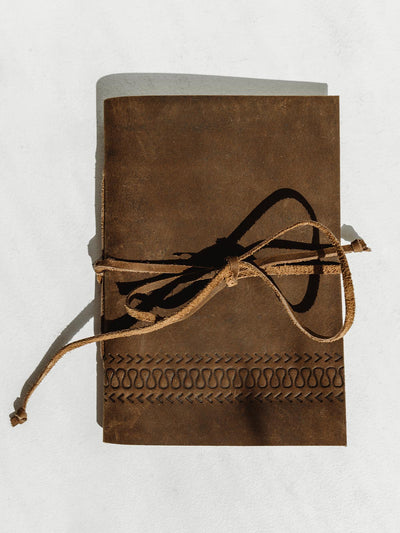 Buffalo Leather journal on a white surface with detail carving at the bottom of the journals cover. Tied closed with a leather strap.