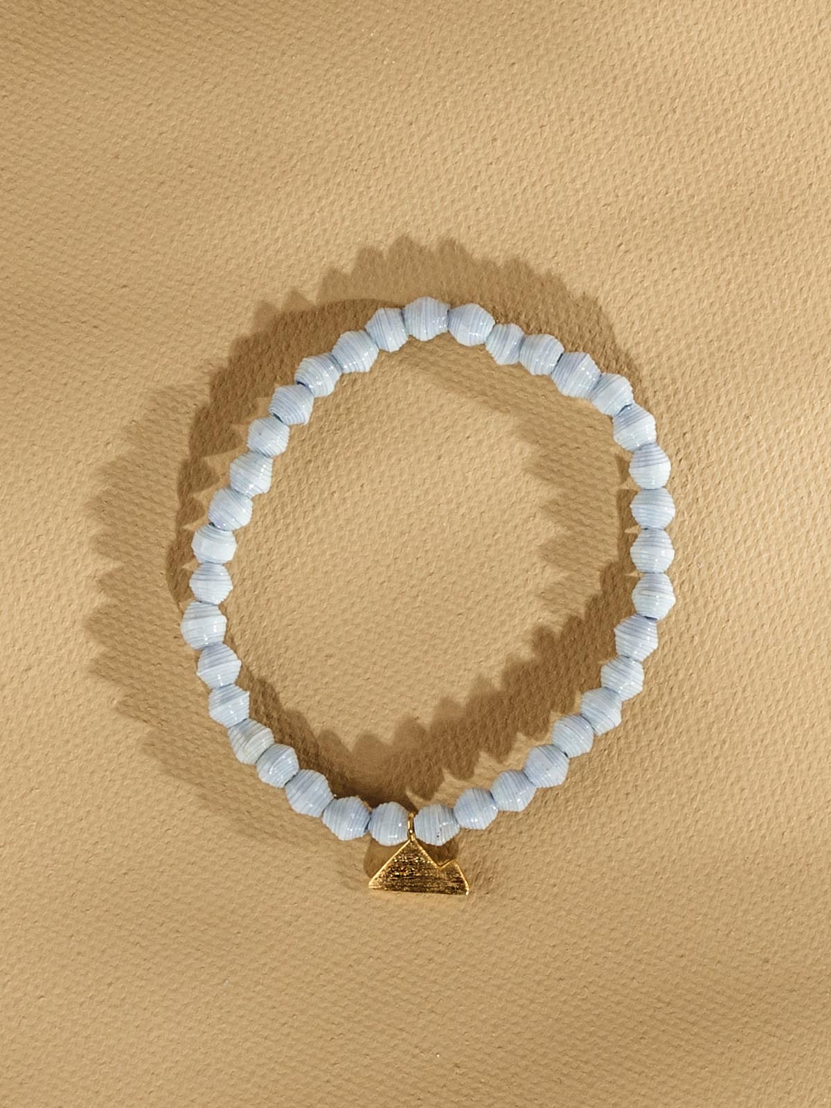 Pale blue beaded bracelet with golden mountain charm. Bracelet is placed on a tan background.