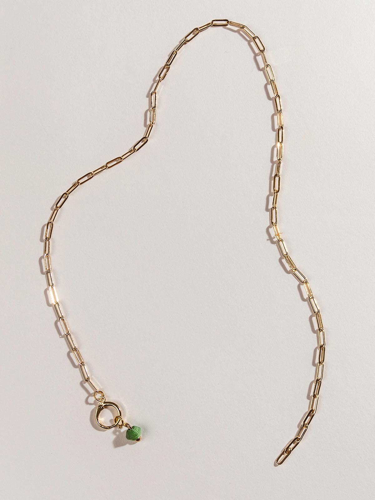 Golden chain charm necklace with one green birthstone bead on the clasp. Necklace is laid on a white surface.