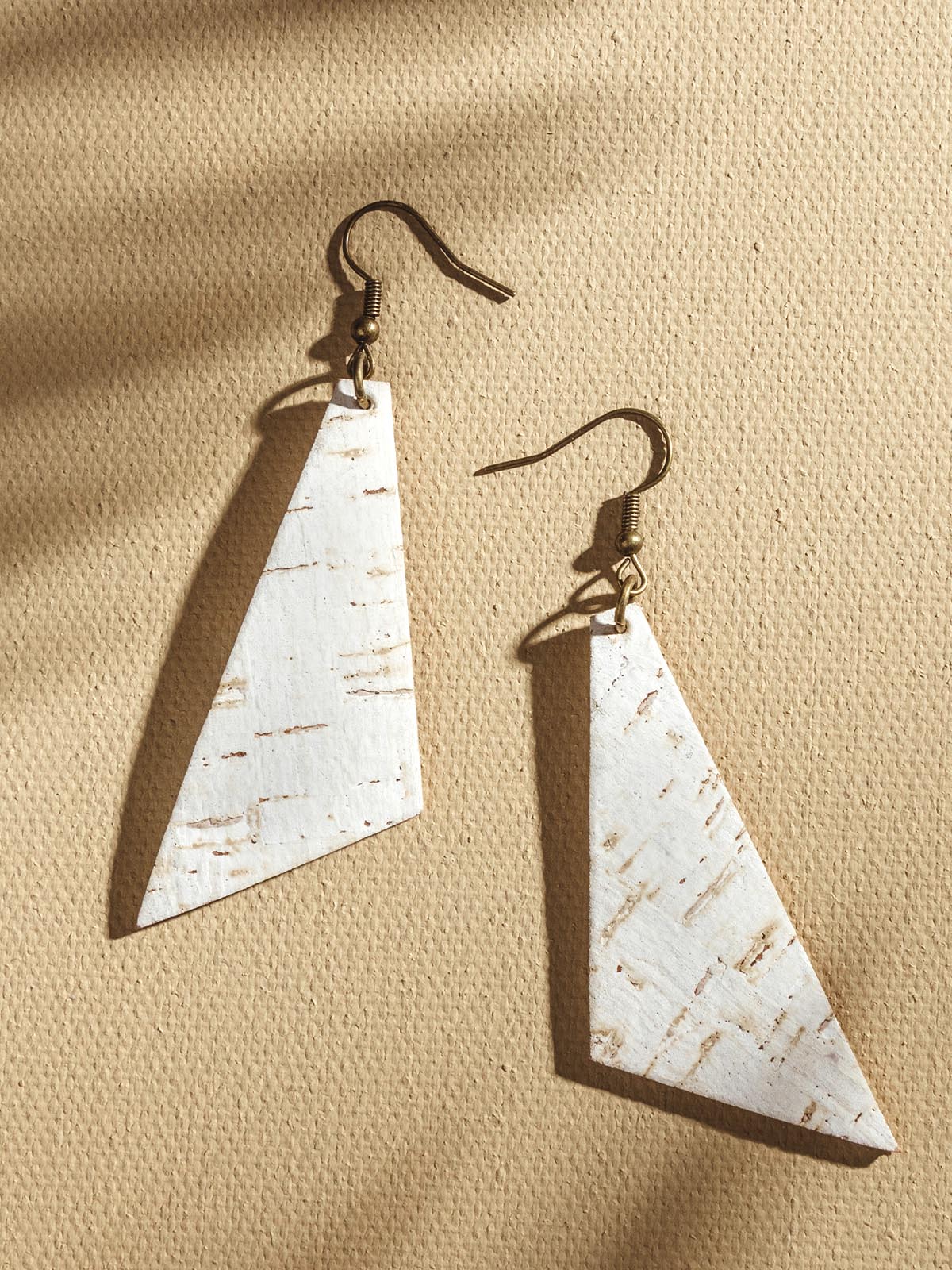 White cork triangular earrings laying on a tan textured background. 