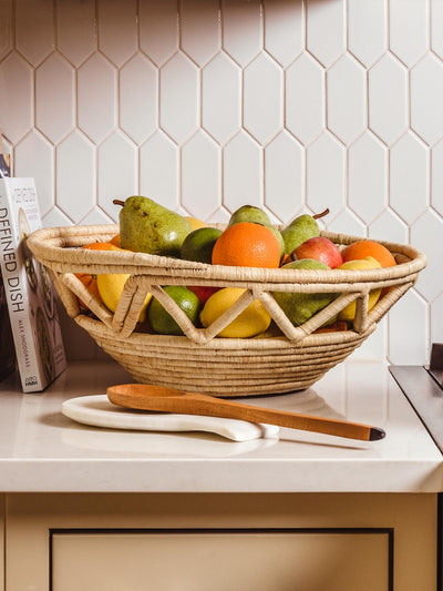Large woven fruit basket full of fruits on kitchen counter with cooking book and wooden spoon.