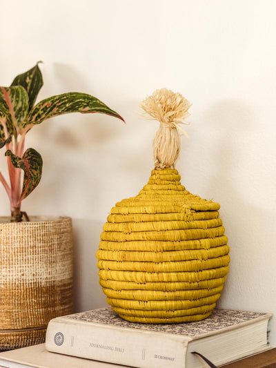 Bright yellow decor basket with tie for opening and closing. Basket is sitting onto of books next to a decorative plant.