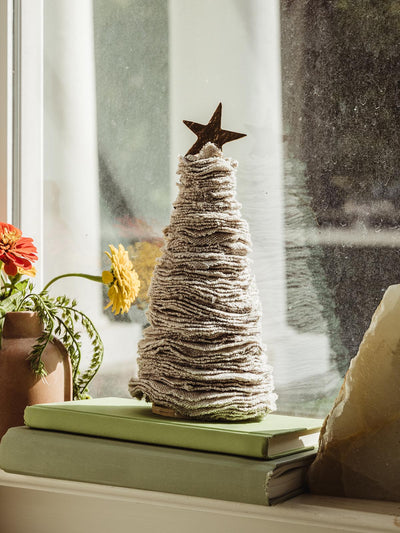 Christmas tree made of cream sweater material sitting in a windowsill onto of books next to flowers and bookends.