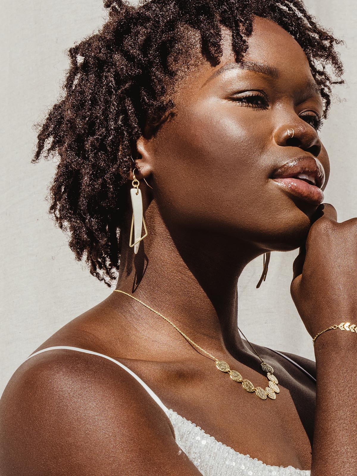Earrings with white triangular dangle layered with thin gold triangular dangle on a female model. Model is wearing other gold jewelry items like necklace and bracelet. 