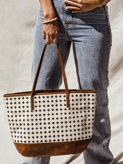 Female model holding large tote with black and white cross pattern with brown leather handle and bottom.
