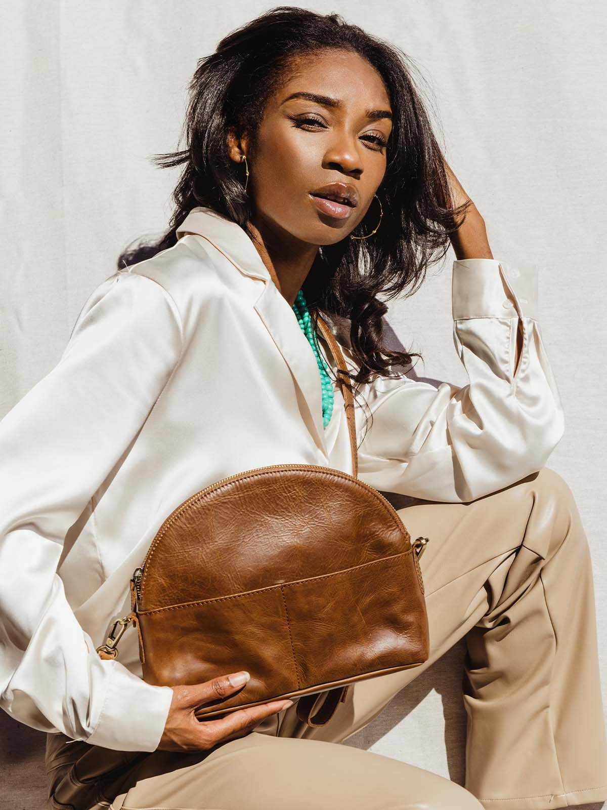 Female model holding brown leather half moon hang bag. Wearing a white silk top and tan leather pants.