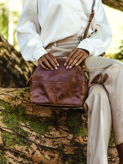 Female model sitting on tree holding half moon leather bag across the body.