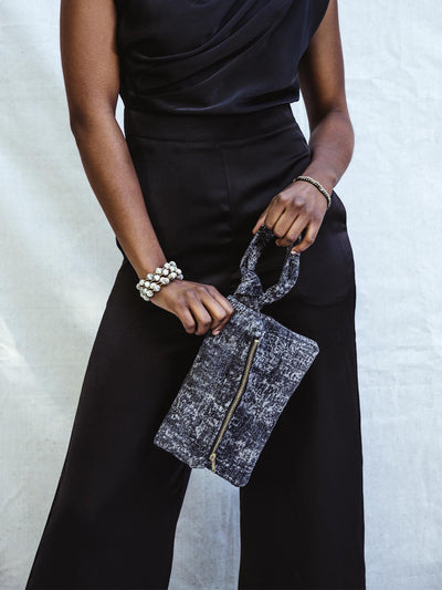 Female model in black silk jumpsuit holding grey cloth cloth with brass zipper and knotted wristlet.