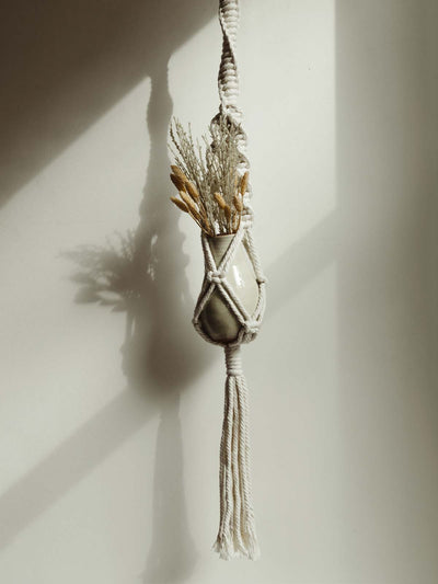 Macrame plant hanging holding vase and dried flowers against a white wall.
