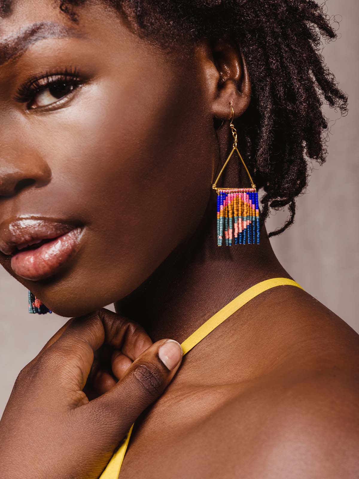 Model wearing golden triangle shape earrings with multi-colored neon beads handing from the triangle shape like fringe. Colors featured are neon pink. blues, and mustard.