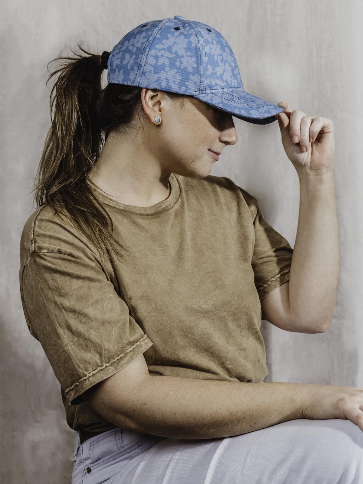 Teal floral hat on girl wearing brown tshirt and blue jeans in front of tan background. 