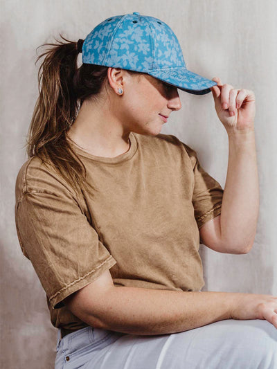Teal floral hat on girl wearing brown tshirt and blue jeans in front of tan background. 