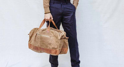 Man wearing black pant with a tan shirt carrying a tan duffle bag