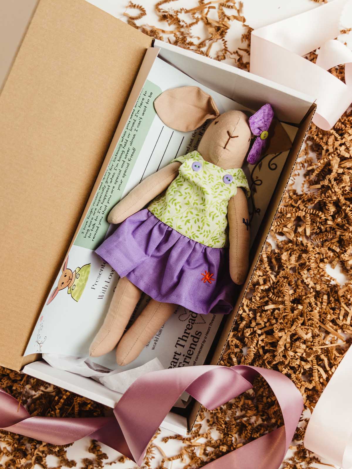 Handmade Heart Thread Friends bunny in a green leaf-print top, purple skirt, and purple bow, nestled in an open gift box on her story card