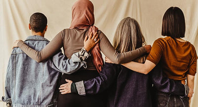 4 women side hugging each other with their backs to the cameras in front of a tan background