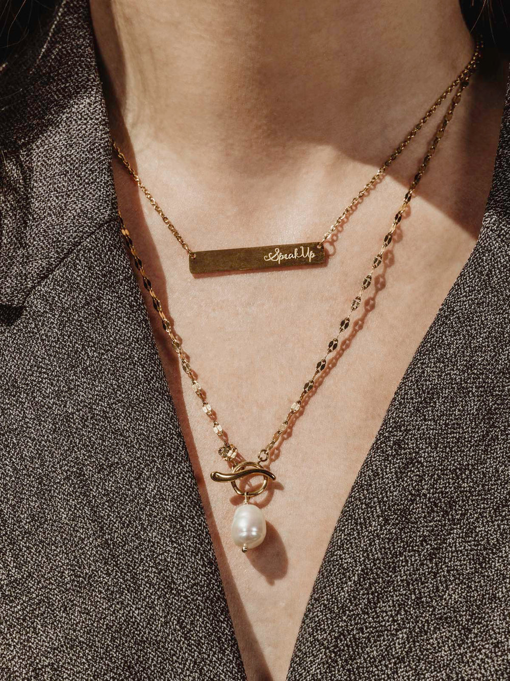 Woman wearing a dark gray blazer with two gold necklaces on. One is gold with the words speak up engraved on it and one is gold chain with a pearl pendent 